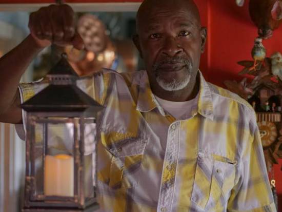 An old black man wearing a short-sleeve. An older man holding a lantern
