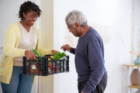 A woman assisting an old man to pick fruit. A young woman holding out a crate of vegetables to her grandfather