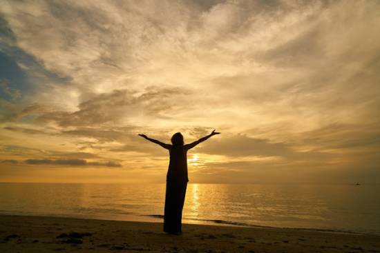 A Praying Woman A silhouette of a woman raising her hands in prayer by the seashore at sunset.