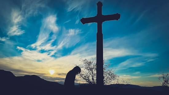 Silhouette of a repentant person kneeling in prayer in front of a cross.
