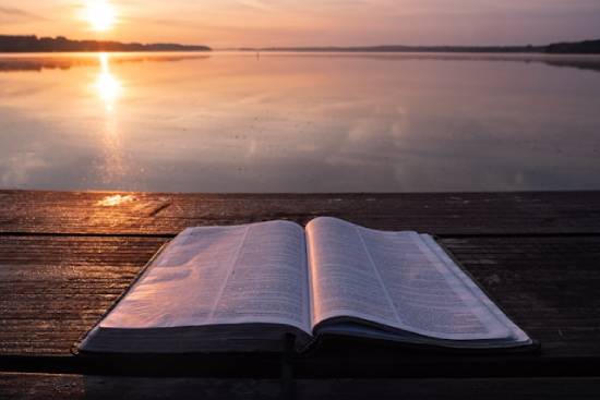 An open Bible on a Table close to a waterbody reflecting on the rays of the setting sun.