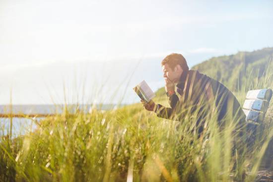 A man sitting on a bench reading the Bible on a grassy lakeshore.