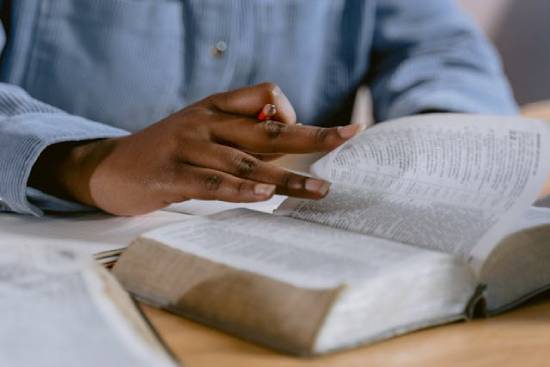 A person in a powder-blue long-sleeved shirt studying the Bible while holding a pencil.
