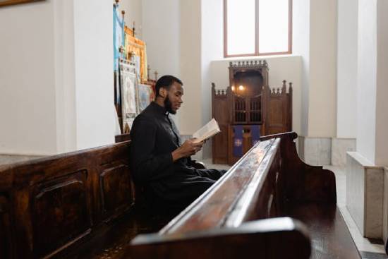 A clergyman in black clerical robe reading the Bible while sitting in the pews.