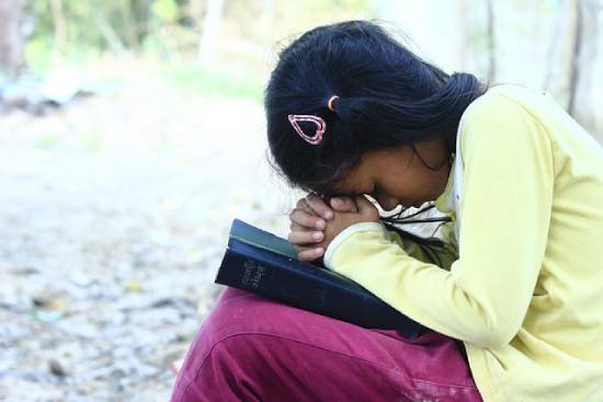 A kid in a mustard jumper and maroon trouser praying with a bible on her kness.