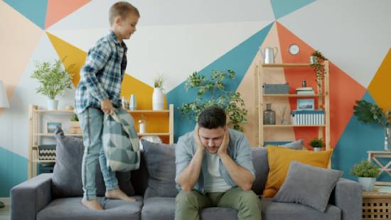 An overwhelmed dad sitting on a couch with palms on his temples as son jumps on the couch.