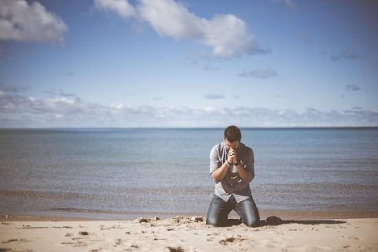 A man kneeling in prayer by the ocean.