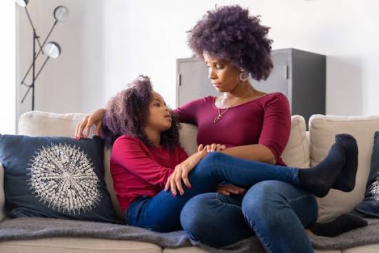 A mother and daughter in burgundy tops and denim trousers having safe conversations while sitting together on a couch.