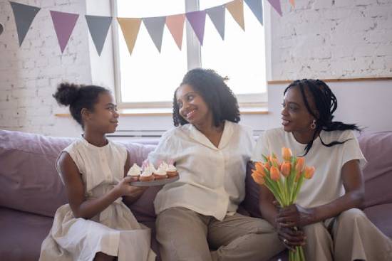 A mother celebrating with two daughters who have brought flowers and cakes.