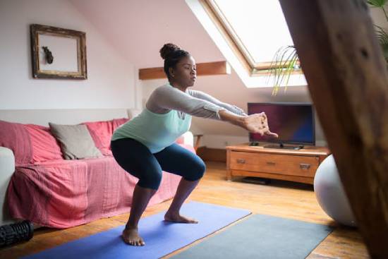 A woman in fitness gear squating while stretching her arms at home.