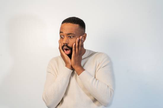 A shocked man in a cream long-sleeved top holding his chin.
