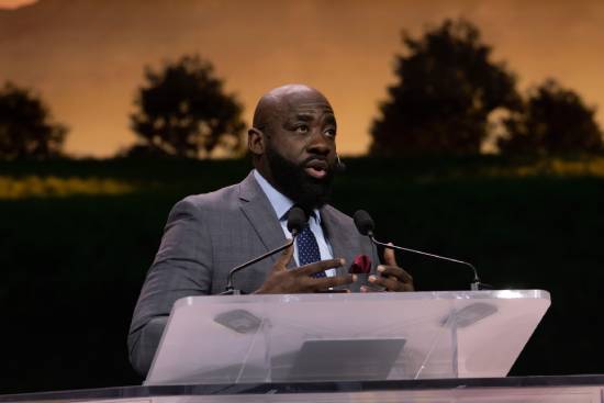 A preacher in a grey suit standing on the pulpit teaching about trusting God in difficult times.