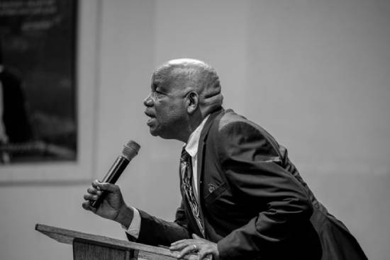 A preacher in a suit talking passionately about faith and emotional growth while leaning on the podium.