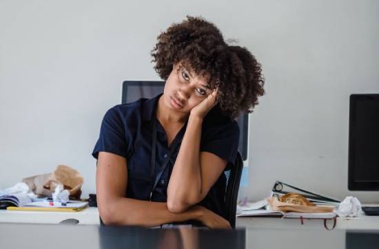 A bored and distracted corporate woman in a blue shortsleebved top sitting on her desk.