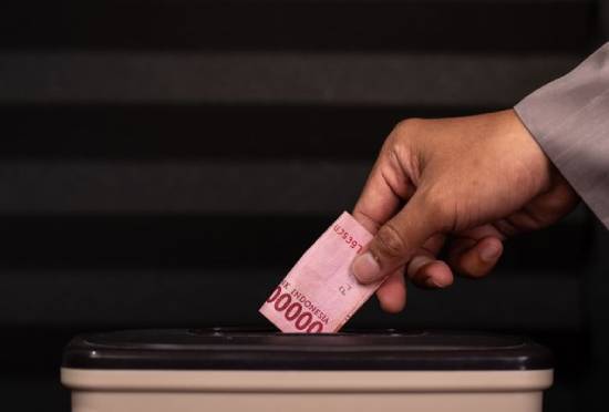 A hand donating Indonesian Rupiah into a donation box.